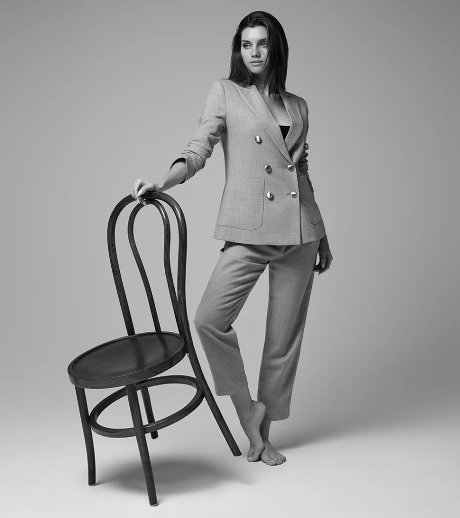 Black and white on-model photography of a double-breasted grey suit, model standing with a bentwood chair