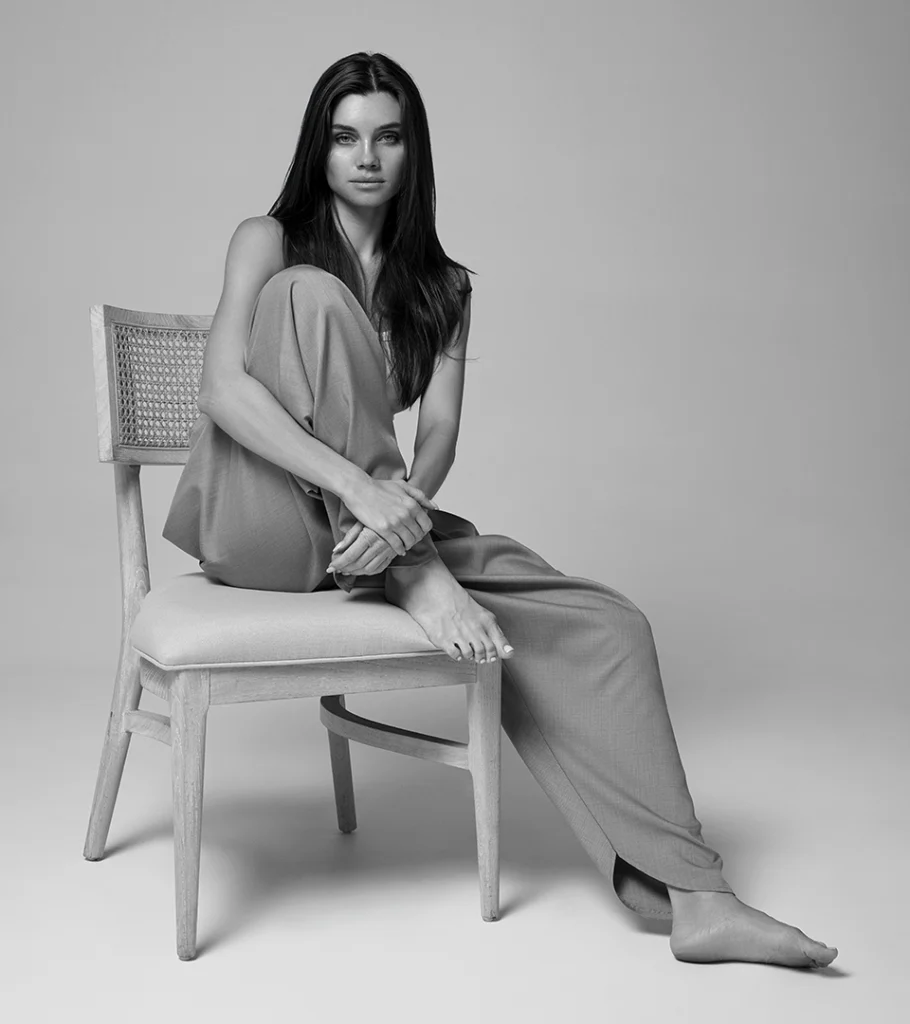 Black and white on-model photography of tailored trousers, model seated barefoot on a chair