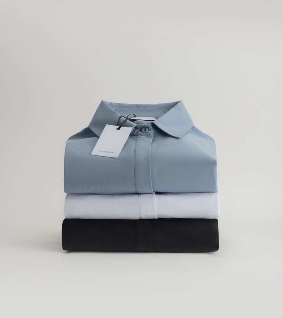 Editorial flat lay photography of three folded shirts stacked in blue, white, and black with a hang tag