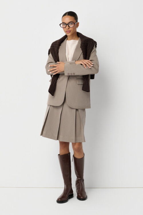 On-model photography of a full taupe blazer and pleated skirt outfit styled with a brown sweater over the shoulders and knee-high boots