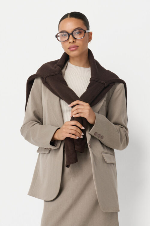 On-model photography of a taupe oversized blazer styled with a brown sweater tied over the shoulders, shown in a three-quarter crop