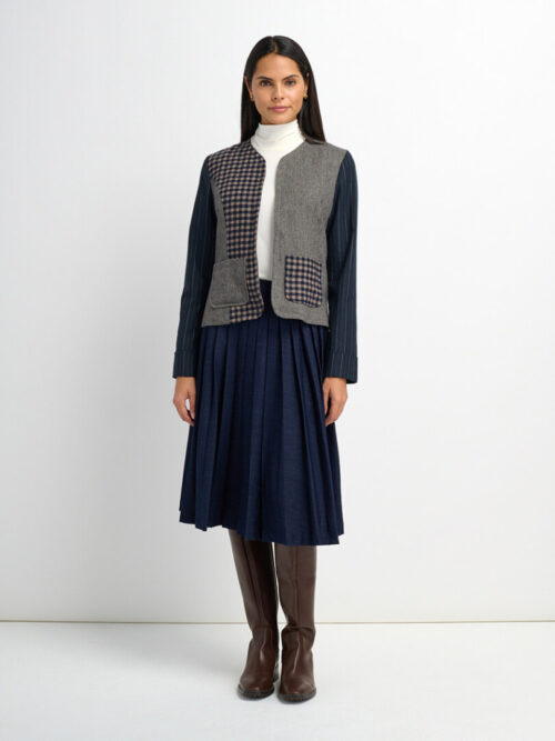 On-model photography of a patchwork check vest layered over a turtleneck with a navy pleated midi skirt and tall boots