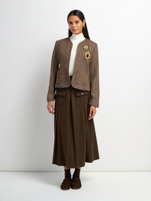 On-model photography of a brown tweed jacket with crest patches worn over a turtleneck with a pleated maxi skirt