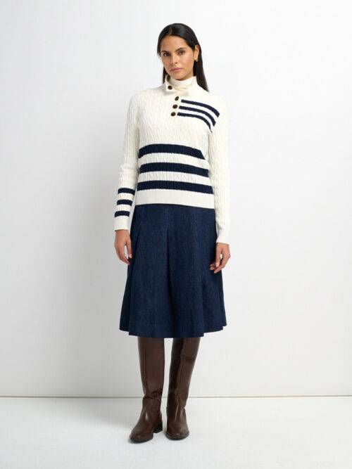 On-model photography of a cream cable-knit sweater with navy stripes and button shoulder, worn with a denim pleated skirt
