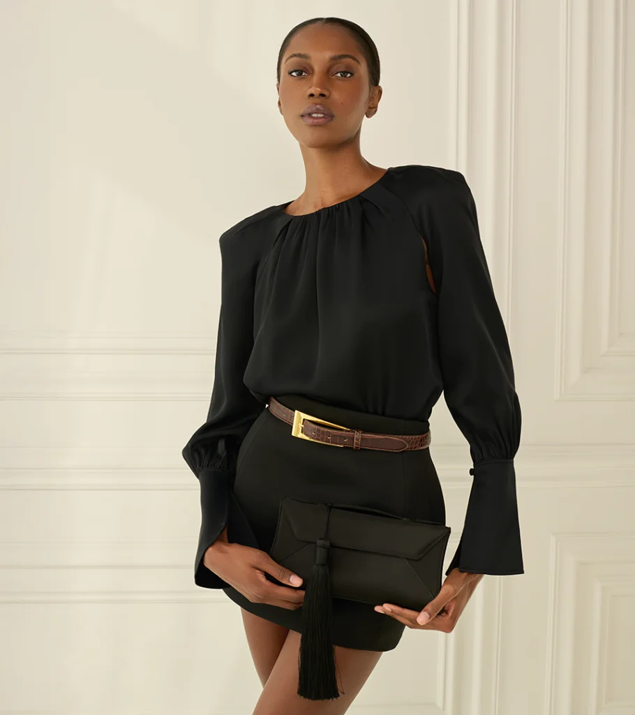 On-model photography of a black long-sleeve blouse and mini skirt with leather belt and tassel clutch