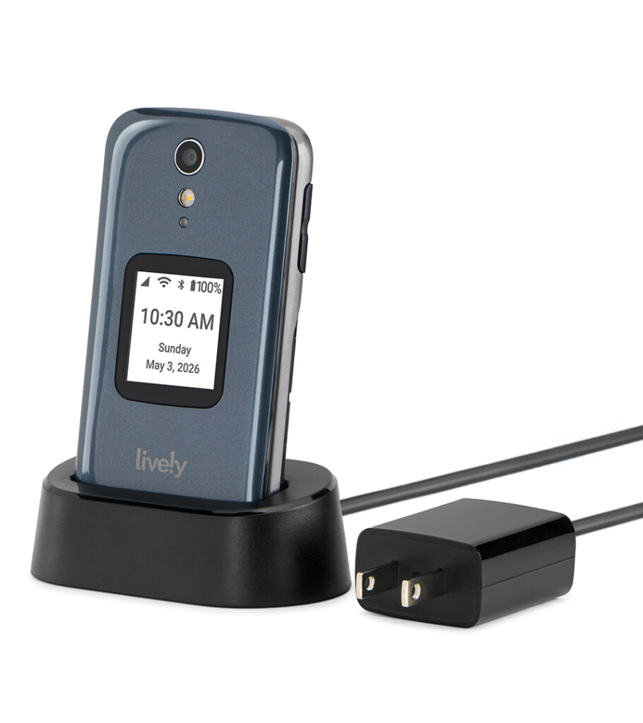 Product photography of a graphite flip phone standing upright in a black charging cradle with the power adapter beside it