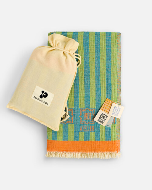 Product photography of a cream drawstring pouch resting on a folded green and blue striped towel with an orange border and fringe