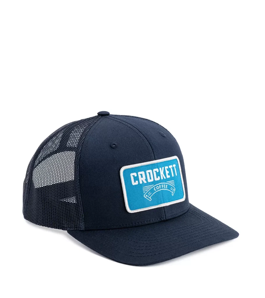 Product photography of a navy mesh-back trucker cap with a blue woven front patch, side view