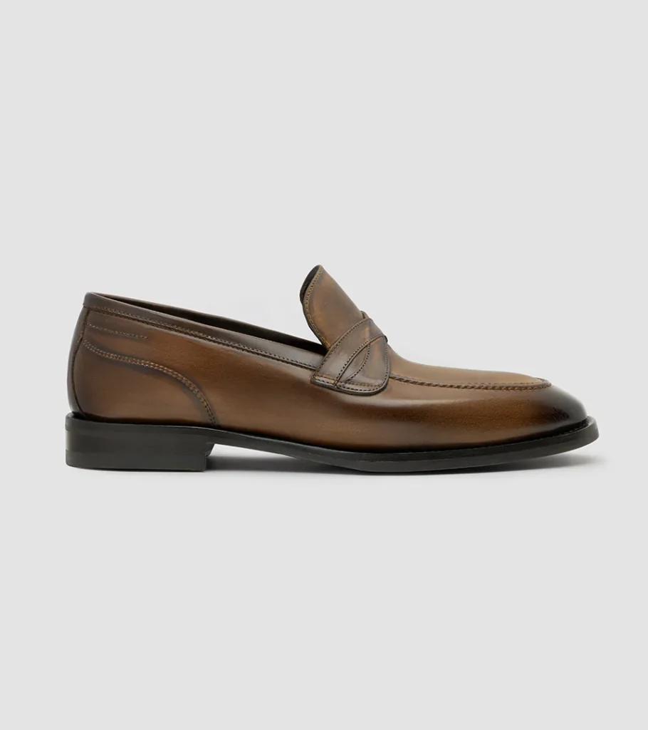 Shoe product photography of a burnished mid-brown penny loafer in side profile showing the saddle strap