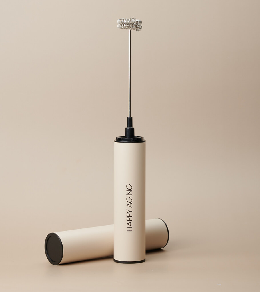 Product photography of a cream handheld milk frother lying beside its matching storage tube, on a warm neutral background