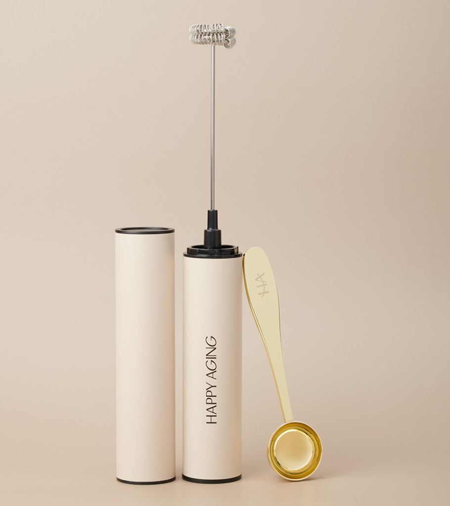 Product photography of a cream handheld milk frother standing upright with its storage tube and a yellow wire whisk attachment