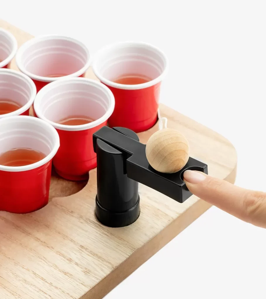 Lifestyle product photography of a hand loading a wooden ball into a tabletop game launcher beside filled red cups