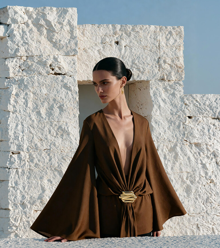 fashion model in brown wide-sleeve blouse with gold clasp posed against white stone wall outdoors