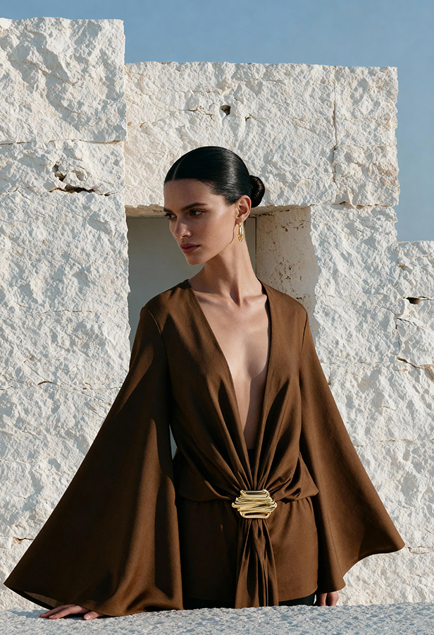 AI-generated models fashion ecommerce by Shootify. Fashion model in a brown bell-sleeve plunge blouse with gold clasp posed against a textured white stone wall.