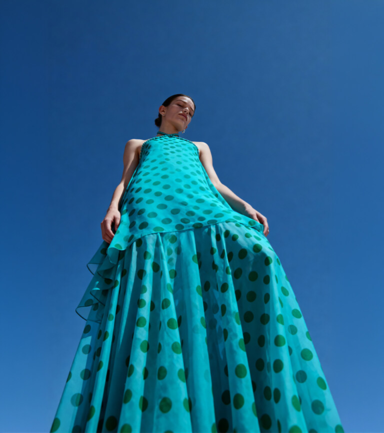 fashion model in teal polka-dot gown photographed from low angle against bright blue sky