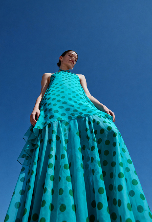 AI models for clothing brands product photography studio. Fashion model in a teal polka-dot tiered halter gown photographed from a low angle against a clear blue sky.