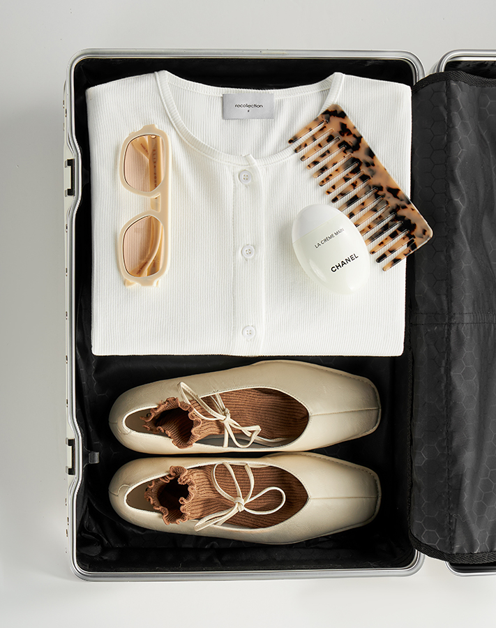 Clothing flat lay and styled product photography by Shootify. Open travel case with white cardigan, cream ballet flats and accessories.