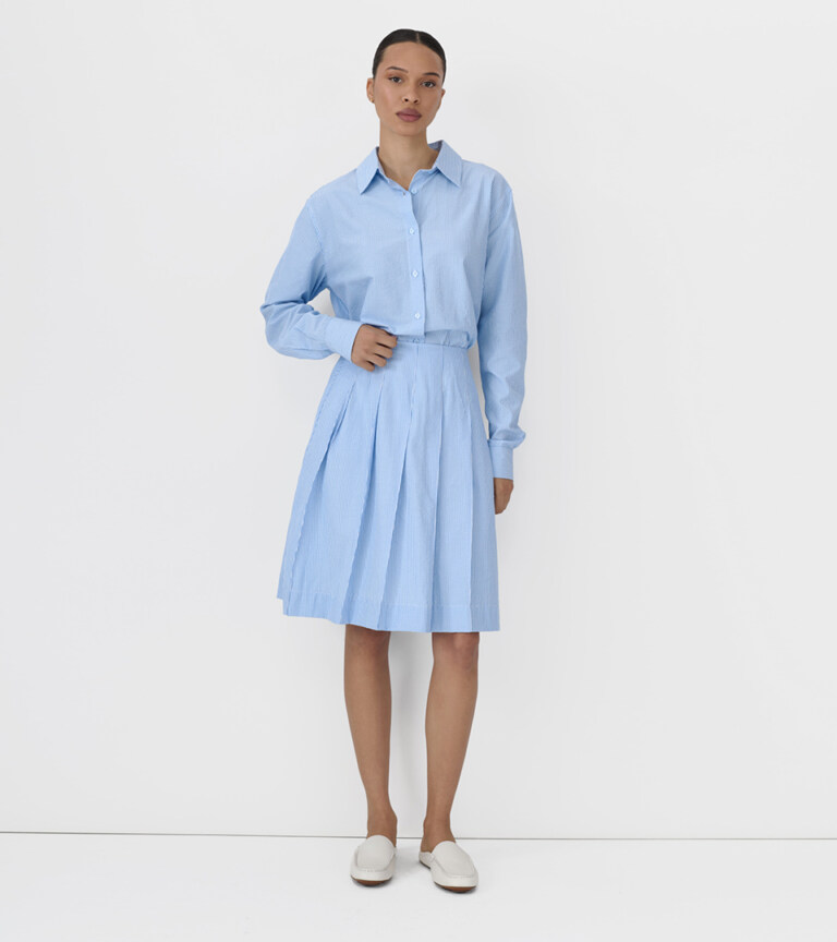 fashion model in blue striped button-up shirt and pleated skirt on white studio background