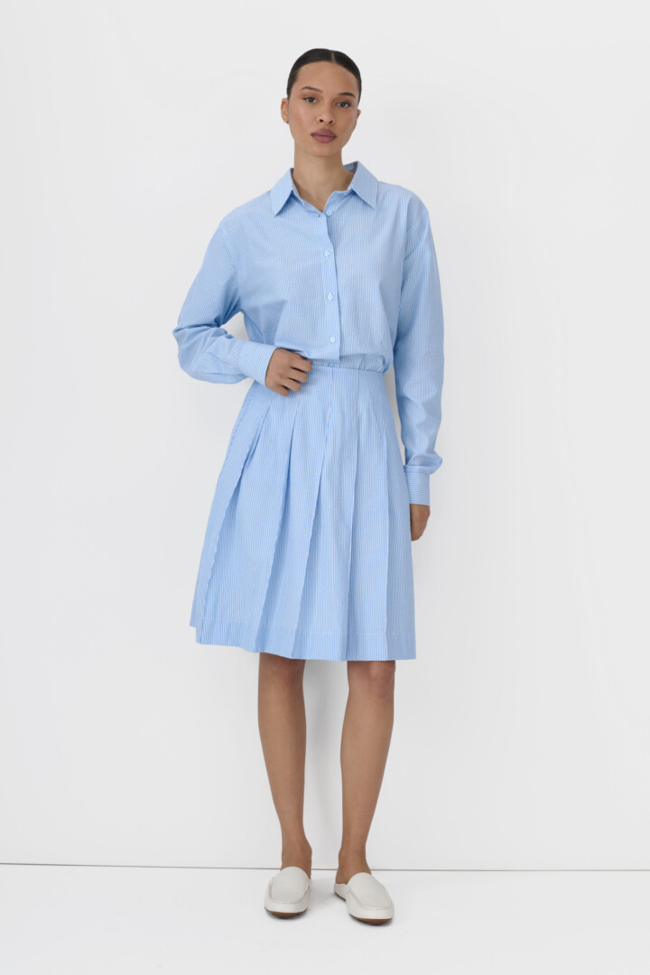 Clothing product video services for ecommerce brands. Model in light blue pinstripe shirt and matching pleated skirt on white.