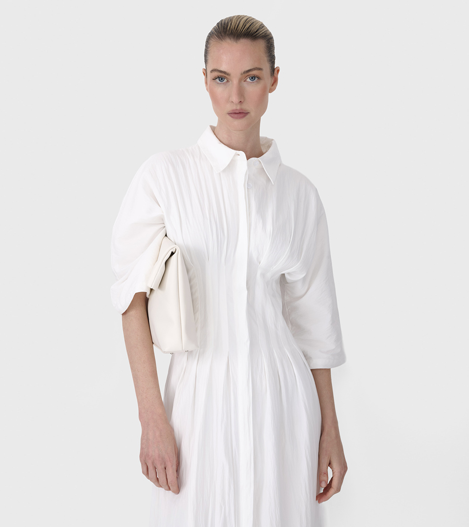 close-up of fashion model in white pleated shirtdress holding cream bag on grey background