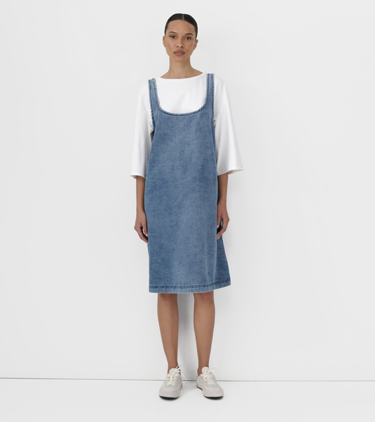fashion model wearing denim pinafore dress over white tee on light grey studio background