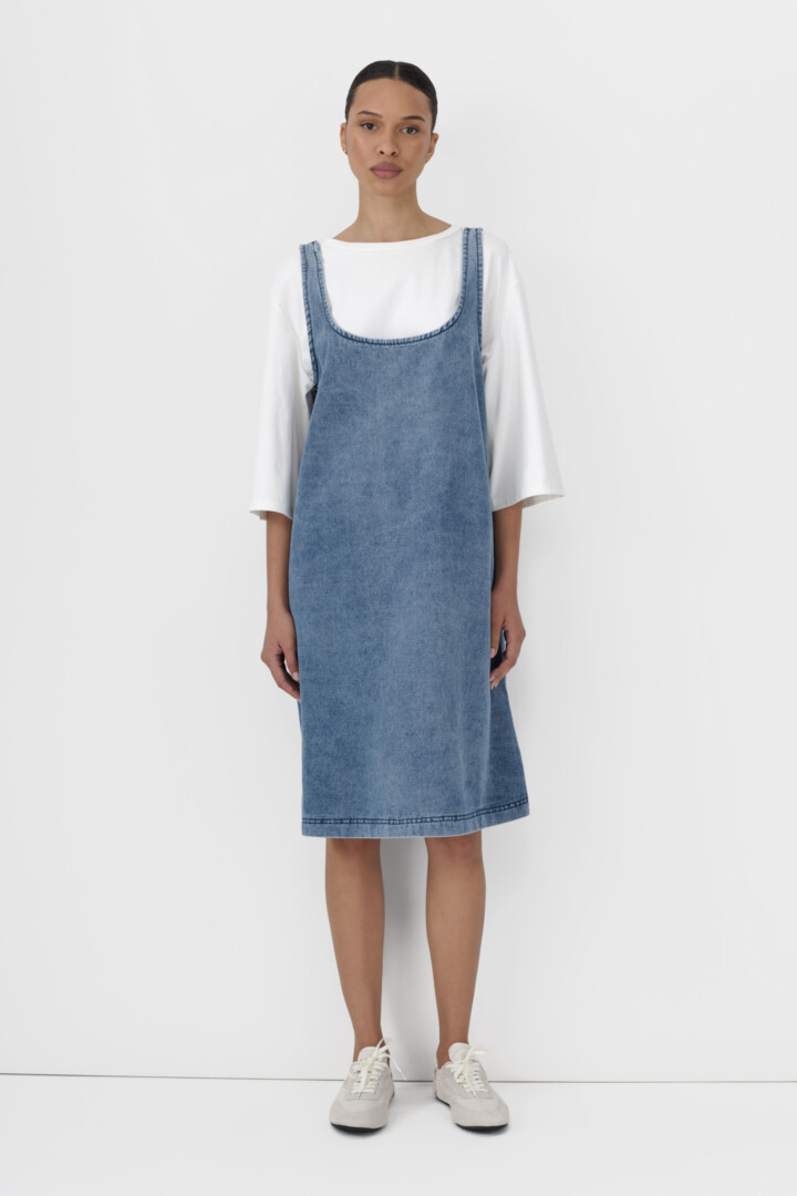 Product photography and video for Shopify stores. Model in light blue denim pinafore dress over white tee on white.