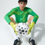 Male model in green goalkeeper kit with gloves, seated, holding a soccer ball