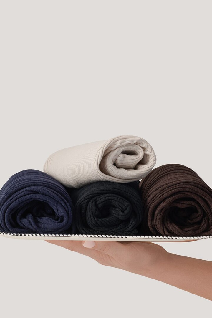 Styled product photography of four rolled knit sweaters in navy, black, cream and brown presented on a white tray held by a hand