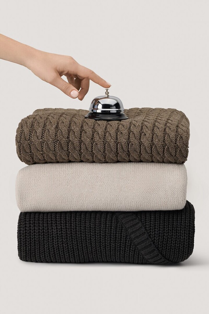 Styled product photography of three folded knit sweaters in brown, cream and black with a hand reaching for a service bell placed on top