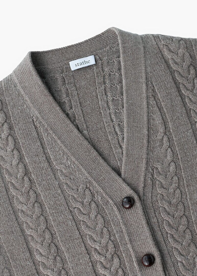 Macro detail photography of a grey cable-knit cardigan showing the cable pattern, ribbed placket, woven brand label and dark horn buttons