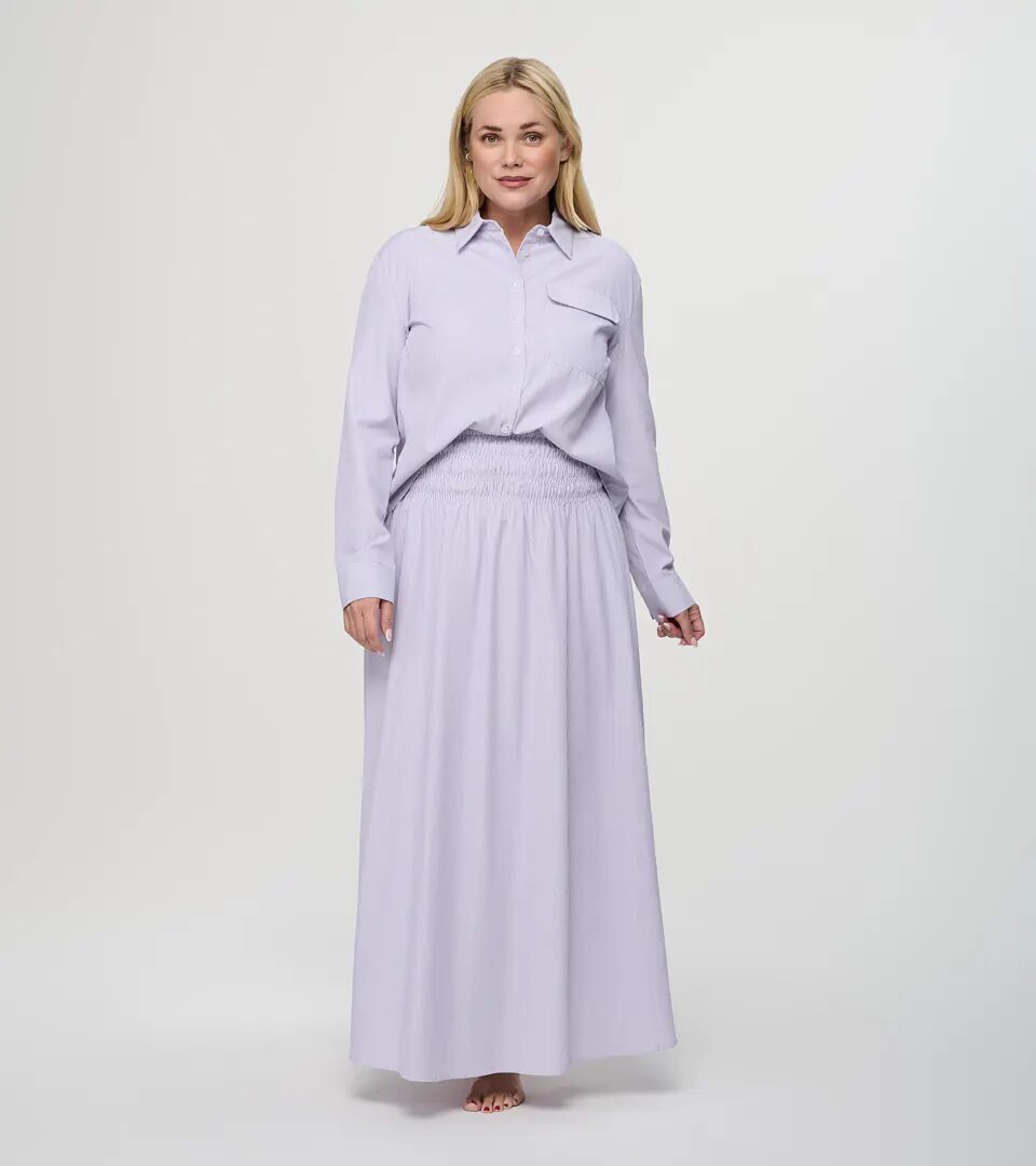 On-model photography of a lilac button-down shirt worn with a matching smocked-waist maxi skirt, full-length front view on a light grey background