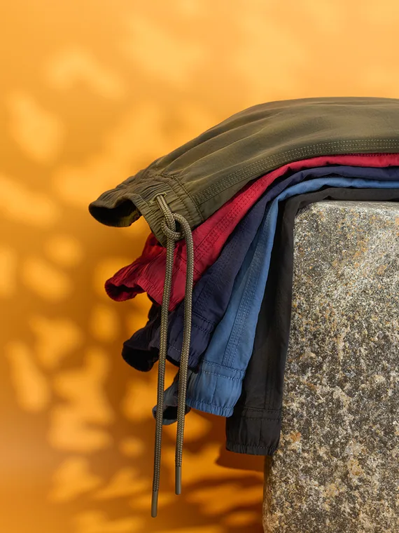 Men's swim trunks stacked on a stone plinth against an orange background with dappled light
