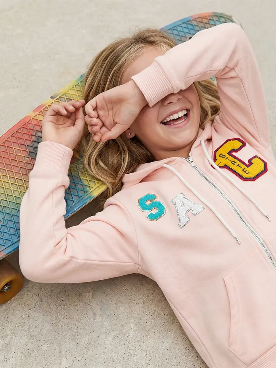 AI model photography of a girl in a pink zip hoodie with varsity letter patches, lying on a skateboard