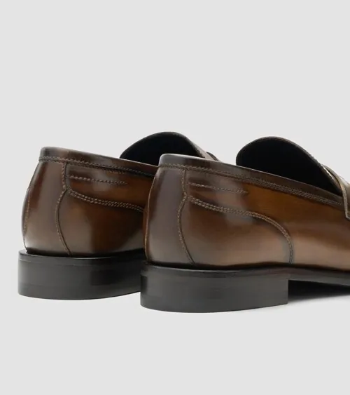Pair of brown burnished leather penny loafers from the back at three-quarter angle on grey background, studio product photography