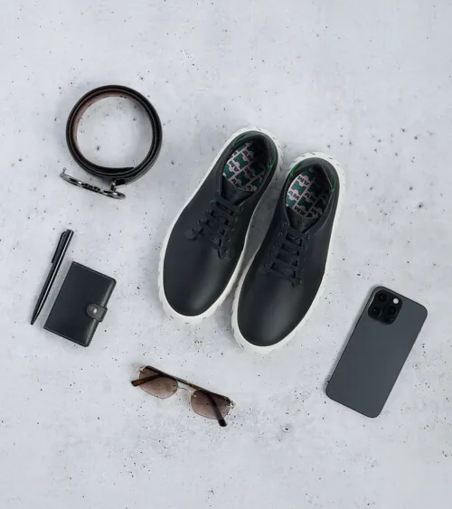 Black sneakers styled flat lay with a belt, wallet, pen, sunglasses, and phone on concrete, lifestyle product photography