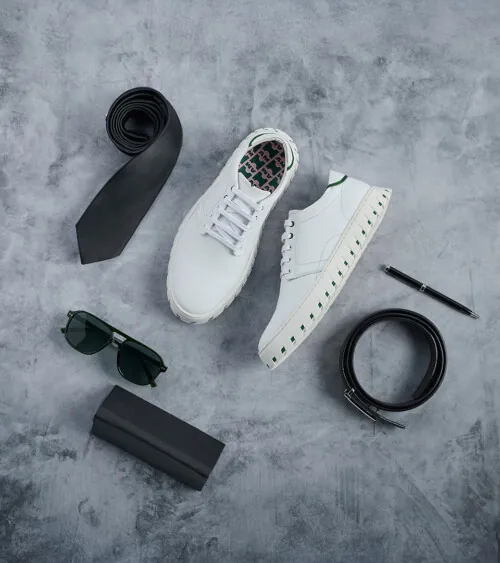 White sneaker styled flat lay with a tie, sunglasses, belt, pen, and wallet on a grey surface, lifestyle product photography