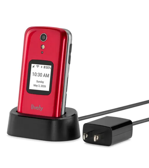 Product photography of a red flip phone standing upright in a black charging cradle with the power adapter beside it