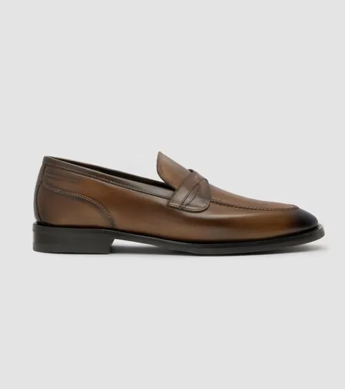 Single brown burnished leather penny loafer shown in side profile on grey background, studio product photography