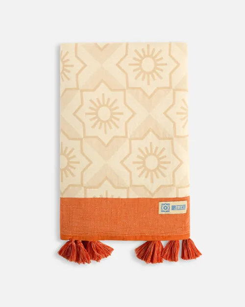 Product photography of a folded cream towel with a floral medallion weave and a terracotta band with tassels