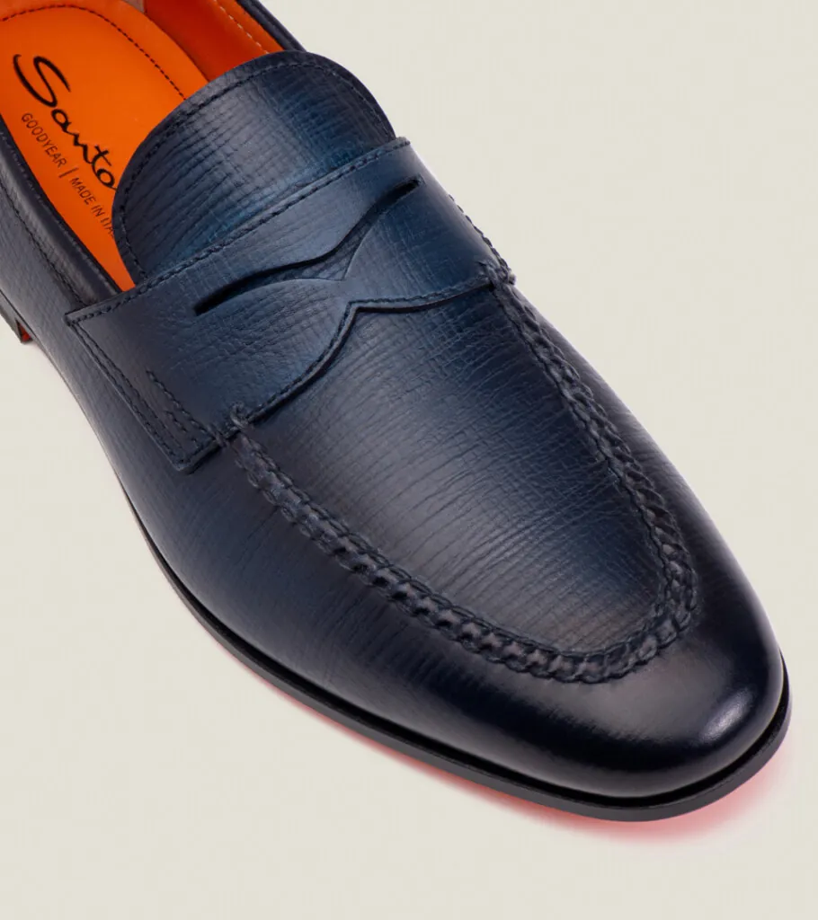 Shoe product photography of a navy penny loafer in close-up at a raised angle showing the saddle strap and stitching